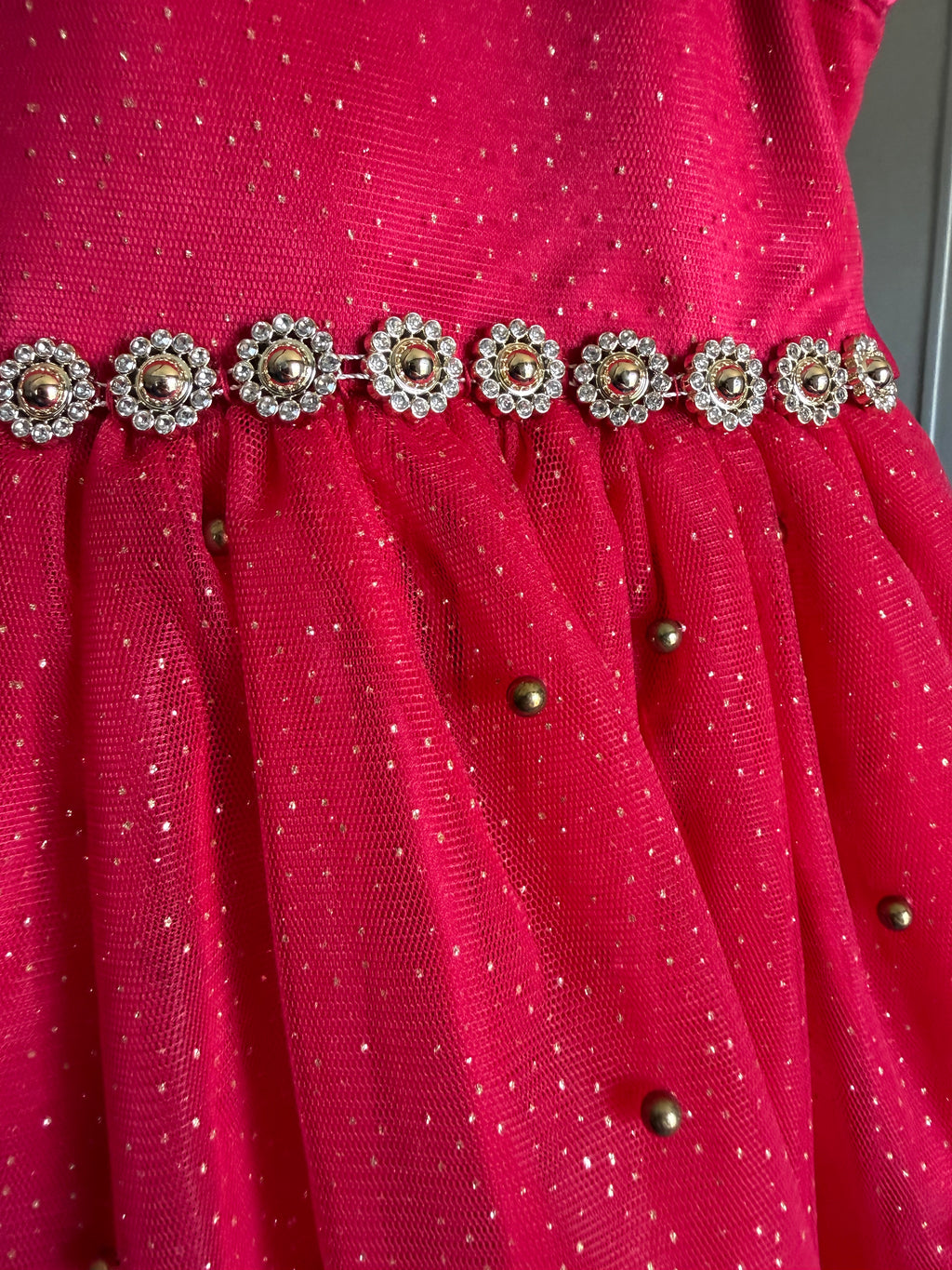 Close-up of a bright red sheer mesh fabric with small metallic gold dots, a decorative waistband of jeweled floral embellishments, and scattered gold metallic bead accents.