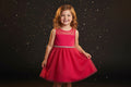 A bright pink sparkly sleeveless dress with a sheer mesh bodice, a gold beaded waistband, and a tulle skirt, hanging on a black hanger in a home setting.